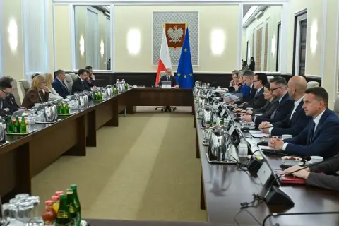 Uczestnicy spotkania przy długim stole, dyskutują o budżetowym deficycie. Na stole napoje i dokumenty.