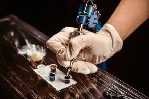Ein Mann bekommt ein Knie Tattoo. Die Tätowiermaschine taucht in schwarze Tinte.
