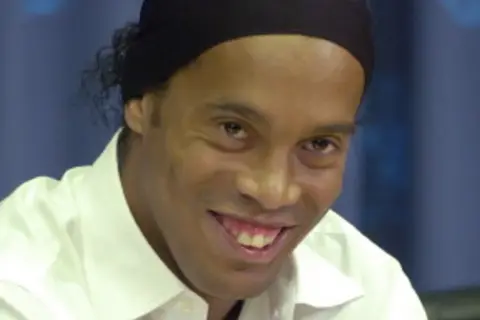 Uśmiechnięty Ronaldinho w białej koszuli i czarnej opasce na głowie, wspominający swoje najlepsze lata w różnych klubach.