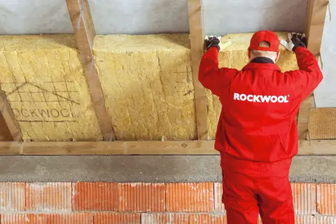 Pracownik w czerwonym stroju Rockwool montuje wełnę mineralną, wykonując ocieplenie murłaty od wewnątrz.