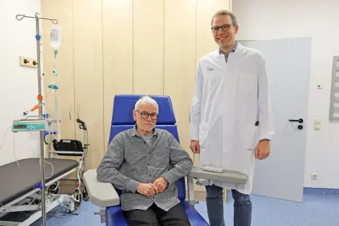 Ein Arzt steht neben einem Patienten, der für eine Alzheimer Behandlung in einem Behandlungsstuhl sitzt. Infusionsständer und medizinische Geräte sind im Hintergrund sichtbar.