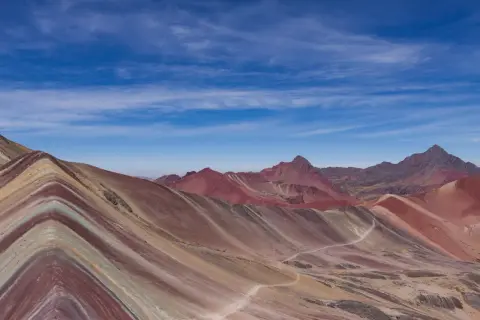 La montagne Vinicunca, aux couleurs vives, s'étend sous un ciel bleu parsemé de nuages. Un sentier serpente à travers ses flancs striés.