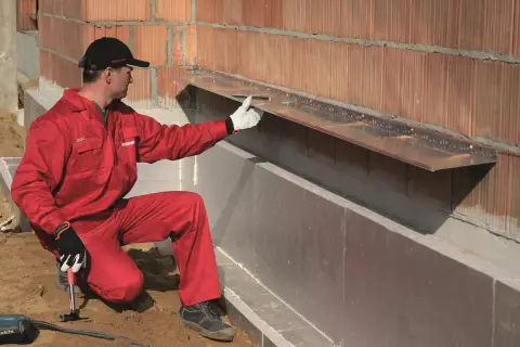 Pracownik w czerwonym kombinezonie montuje okapnik na ścianie zewnętrznej, przygotowując ją do montażu wełny mineralnej.
