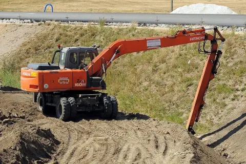 Pomarańczowa koparka Hitachi Zaxis 160W pracuje na budowie. Operator posiada odpowiednie uprawnienia na koparki, by sprawnie wykonywać prace ziemne.
