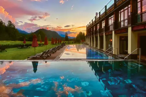 Abendrot über den Alpen spiegelt sich im Infinity-Pool eines der besonderen Hotels Bayern. Liegestühle und rote Sonnenschirme säumen den Rasen.