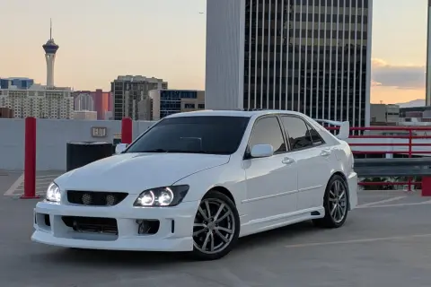 Biała Toyota Altezza z tuningowym body kitem i spojlerem na dachu parkingu, w tle wieżowce i wieża widokowa.