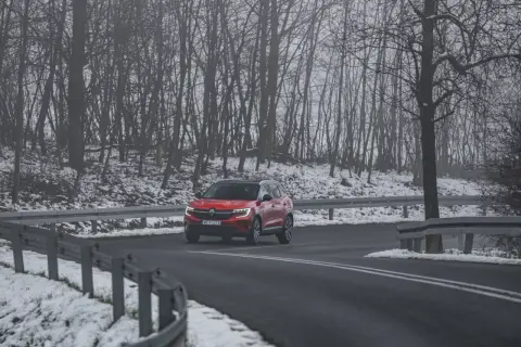 Czerwony SUV jedzie po mokrej drodze, otoczonej ośnieżonym lasem. Kierowca uważa, bo to może być gololedz.
