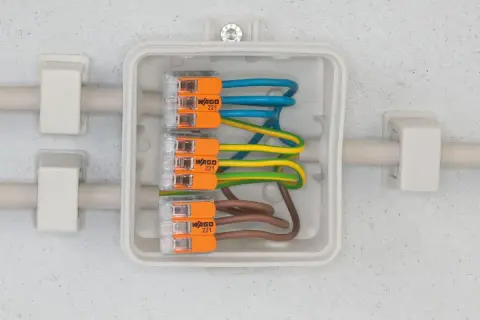 Nowoczesna instalacja elektryczna z kostkami WAGO 221, łączącymi przewody niebieski, żółto-zielony i brązowy w białej puszce.