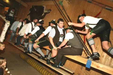 Männer in Lederhosen und Trachtenhemden auf einer Holzbank, bereit für ein Spiel auf der bayrischen Kirchweih.