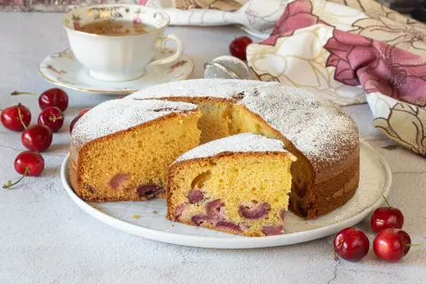 Una fetta di torta alle ciliegie, soffice e profumata, cosparsa di zucchero a velo. Ciliegie fresche e una tazza di tè completano la scena.