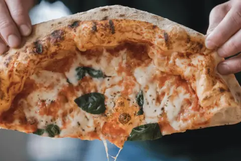 Hände halten ein Stück Pizza mit luftigem Rand, geschmolzenem Käse und Basilikum.