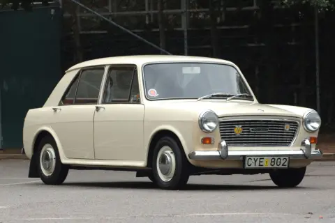 En krämvit Austin 1100 står parkerad på asfalt. Bilen har en klassisk design med kromade detaljer och en tydlig grill.