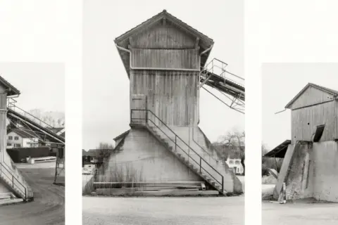 Strutture industriali in legno e cemento, tipiche delle opere di Hilla Becher, con scale esterne e nastri trasportatori.