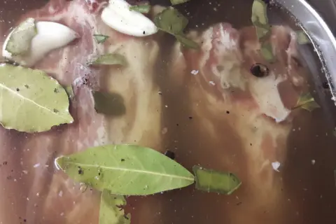 Mięso w mętnej, brązowej zalewie z liśćmi laurowymi i czosnkiem. Wygląda jak zepsuta solanka, pełna drobnych zanieczyszczeń.