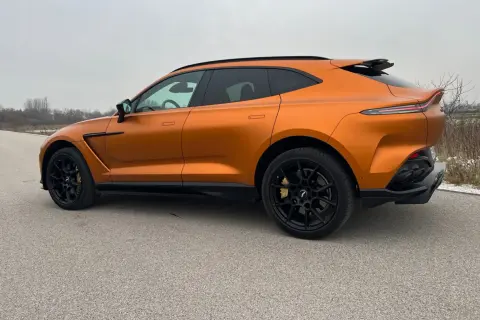 Pomarańczowy Aston Martin DBX z czarnymi felgami i przyciemnianymi szybami na tle pochmurnego nieba.