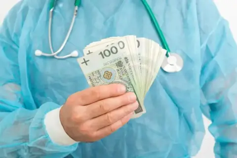 Lekarz rezydent w niebieskim fartuchu trzyma w dłoni plik banknotów 100 zł. Pytanie brzmi: ile zarabia lekarz rezydent?