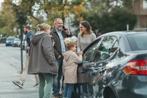 Familie am Auto, Vater telefoniert, vielleicht wegen eines Problems, das die Unfallkasse des Bundes betrifft.