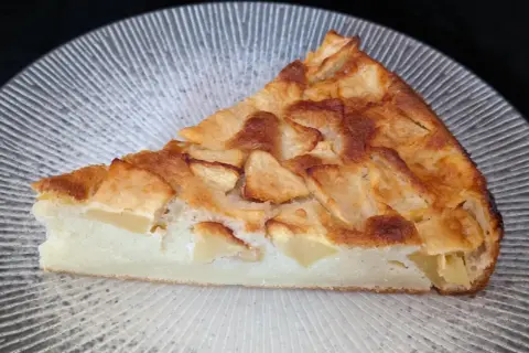 Tranche de clafoutis aux pommes dorée et moelleuse, garnie de morceaux de fruits fondants, sur une assiette texturée.