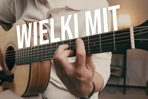 Ręka grająca na gitarze akustycznej, palce na gryfie. W tle biały napis "WIELKI MIT". Idealne ćwiczenia gitarowe.