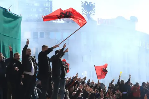Människor viftar med albanska flaggor och gör fredstecken. En del av folkmassan verkar fira, vilket ger en känsla av stolthet och gemenskap. Detta kan vara en del av fakta om Albanien.