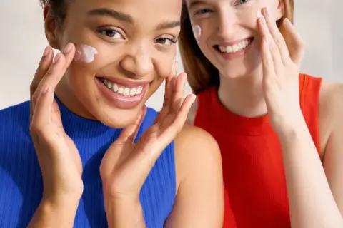 Dos mujeres sonríen mientras se aplican crema en el rostro. Ambas tienen piel normal y lucen radiantes.