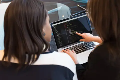Dwie kobiety analizują kod na laptopie. Jedna wskazuje palcem na ekran, druga pisze. Tak wygląda praca programisty.