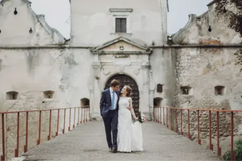 Ein Brautpaar spaziert über eine Brücke vor einer alten Burg. Die Szene erinnert an eine mittelalterliche Hochzeit.
