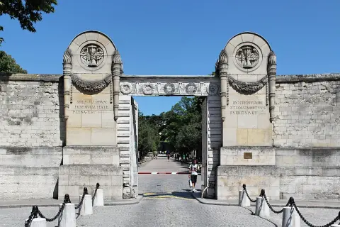 Monumentalna brama cmentarza Père Lachaise z łacińskimi inskrypcjami i płonącymi pochodniami.