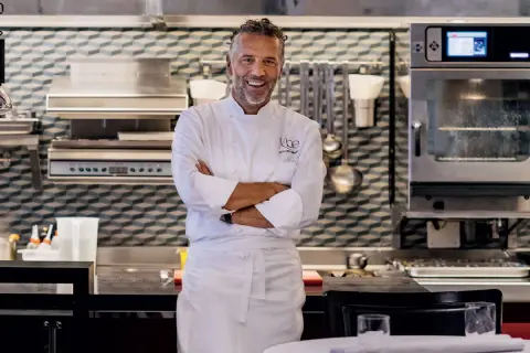 Chef Perbellini, sorridente in cucina, con le braccia conserte. Dietro di lui, attrezzature professionali e un forno acceso.