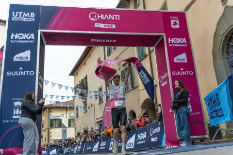 Un coureur franchit la ligne d'arrivée du Chianti by UTMB, acclamé par la foule. Il tient une banderole rose de finisher.