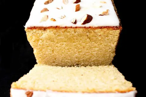A slice of almond cake with white icing and chopped almonds on top, against a black background.