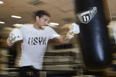 A boxer in a white t-shirt and gloves trains intensely during a boxing training camp, punching a heavy bag.