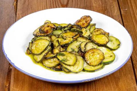 Zucchine alla poverella, fette sottili e dorate, condite con olio e erbe aromatiche, su un piatto bianco con bordo blu.