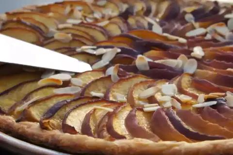 Une tranche de tarte aux nectarines, garnie d'amandes effilées, est coupée par un couteau.