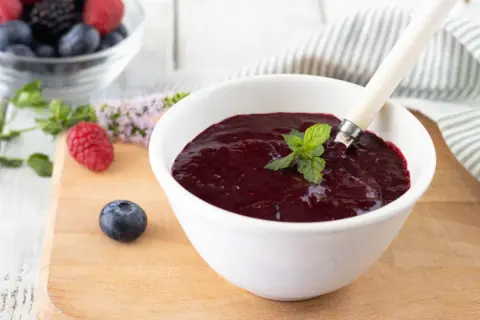 Una ciotola bianca contiene un denso coulis frutti di bosco, guarnito con menta fresca. Frutti di bosco freschi e foglie di menta sono sparsi sul tagliere.