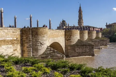 El histórico puente de piedra cruza un río caudaloso. En la orilla, gente pasea y se admira la torre de una iglesia.