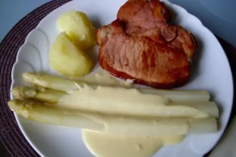 Ein Teller mit gebratenem Schinken, Kartoffeln und Spargel in Sauce Hollandaise. Der Spargel ist in Alufolie gewickelt.