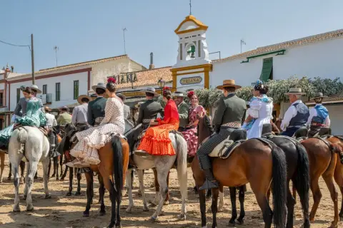 Gente vestida de flamenco a caballo, en un **camino rocío** bajo un cielo azul. Edificios blancos y un campanario al fondo.