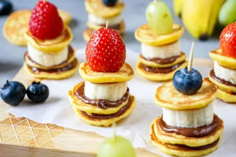 Kleine, gestapelte Baby-Pfannkuchen mit Banane und Schokoaufstrich, garniert mit Beeren und Trauben. Ein süßer Genuss!