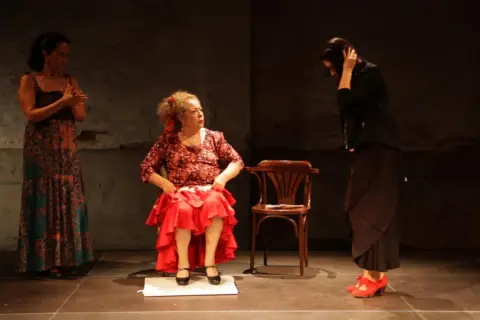 Tres mujeres en escena. Una sentada, con falda roja y blusa estampada, como **la Chana**. Otra de pie, con vestido largo y estampado. La tercera, de negro, con el pelo recogido.