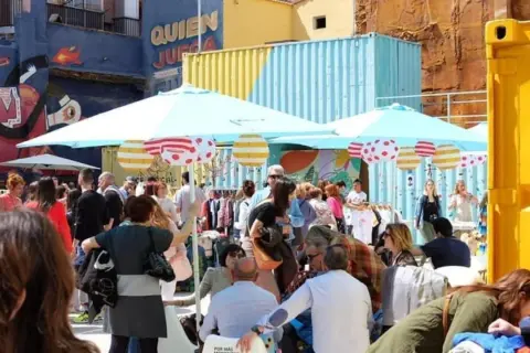 Gente comprando ropa en un animado mercadillo de las armas, con coloridos contenedores y murales de fondo.