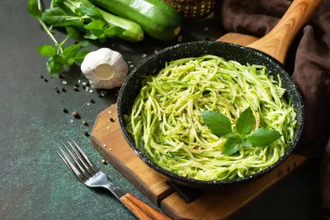 Pyszny makaron keto z cukinii w patelni, udekorowany bazylią. Obok świeże cukinie, czosnek i przyprawy.