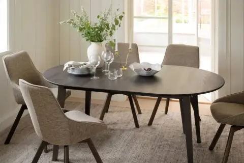 Ett runt mörkt matbord med fyra stoppade stolar runt sig. En vas med blommor, ljusstakar och en skål med frukt på bordet. En tidlös design klassiker.
