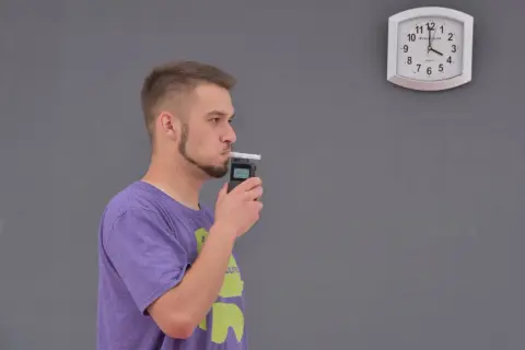 Młody mężczyzna dmucha w alkomat, sprawdzając, ile promili ma po lampce szampana. Zegar na ścianie pokazuje czas.