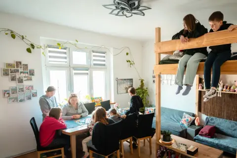 Rodzina gra w planszówkę w domu pomocy społecznej "Przystań". Dzieci siedzą na łóżku piętrowym, obserwując grę.