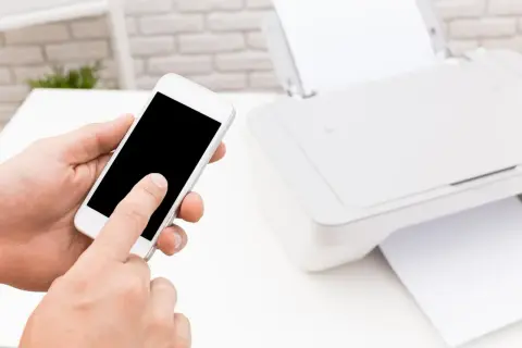 Dłoń trzyma smartfon, inicjując drukowanie z telefonu przez wifi. Obok drukarka z papierem.