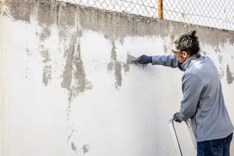 Hombre raspa pared desconchada, preparándola para pintura alquitranada.