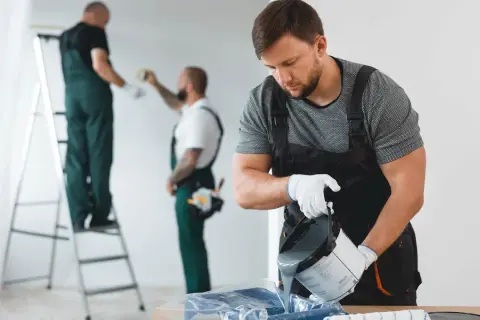 Malowanie ścian w bloku. Dwóch mężczyzn w kombinezonach przygotowuje się do pracy, jeden nalewa farbę, drugi jest na drabinie.