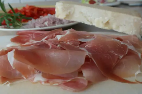 Cieniutkie plastry szynki crudo, obok kawałek parmezanu i talerz z posiekanymi pomidorami i rozmarynem.