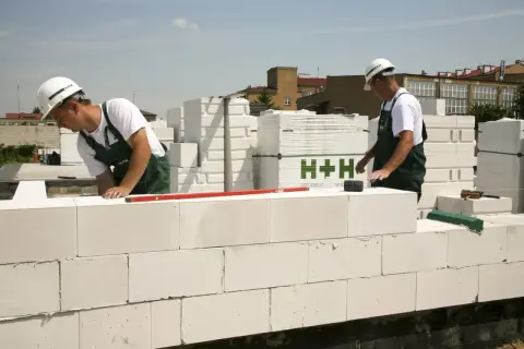 Budowlańcy układają bloczki z **suporeksu**, tworząc ścianę. W tle widać palety z materiałem budowlanym.